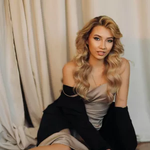 Blonde woman in a glamorous dress posing elegantly indoors against a neutral curtain backdrop.