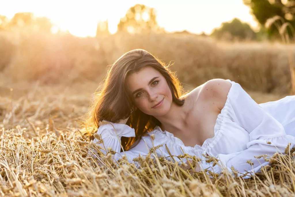 A beautiful woman in a white top lies in a golden wheat field during sunset, smiling softly.
