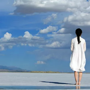 Woman stands alone on a vast salt flat under a bright blue sky with clouds.
