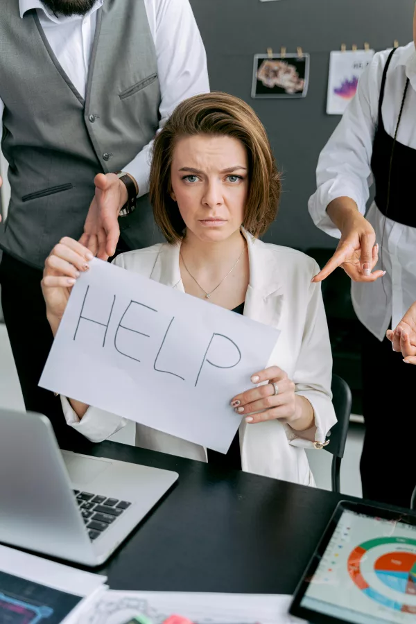 A stressed woman in an office holds a help sign while colleagues point and surround her.