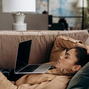 Exhausted woman rests with laptop on sofa, illustrating work-from-home fatigue.