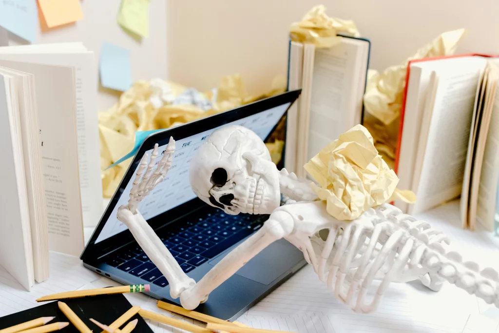 A skeleton surrounded by crumpled paper and books at a messy desk symbolizes burnout and overwork.