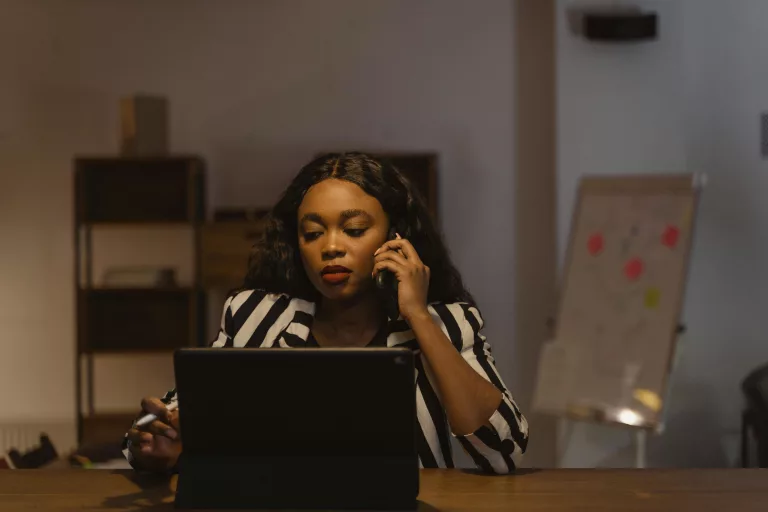 Focused businesswoman in striped blazer working at office desk on tablet while on a phone call.