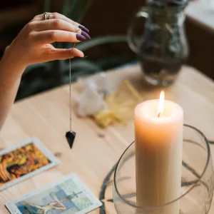 From above of crop anonymous female fortune teller with amulet on thread above tarot cards near burning candle on table